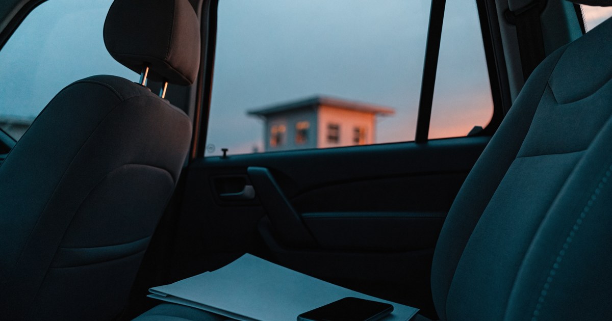 A car passenger seat at dusk with a folded paper folder and a phone, with a small softly lit building outside the window.