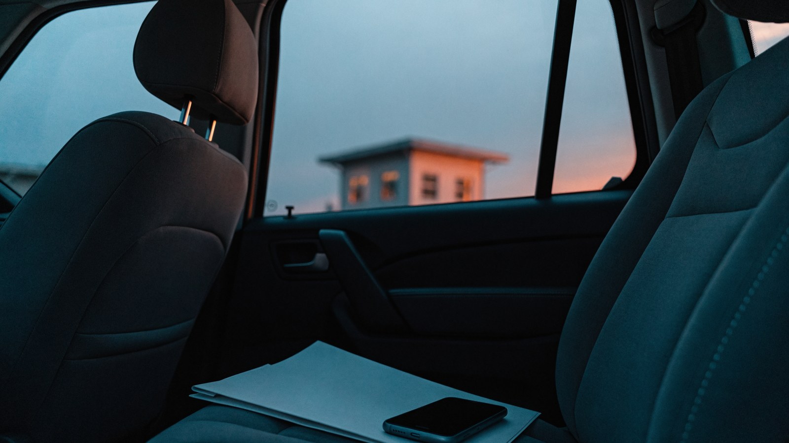 A car passenger seat at dusk with a folded paper folder and a phone, with a small softly lit building outside the window.